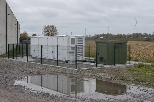 Batterijcontainers achter een omheining naast een bedrijfshal, met windmolens in de verte