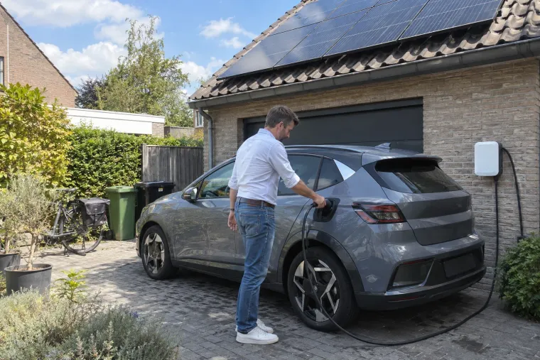 Man steekt overdag de laadkabel in zijn wagen, onder de zonnepanelen op de garage